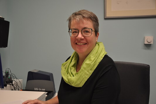 Frau mit Brille und grünem Schal sitzt lächelnd am Schreibtisch im modernen Büro.